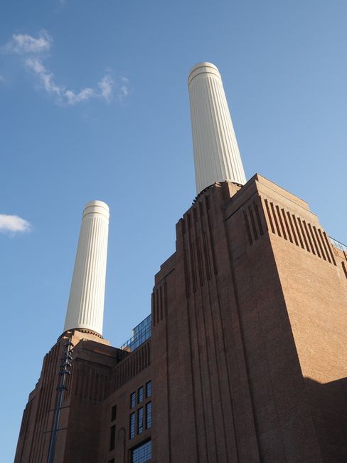 A large, historic power station building made of red brick with tall, cylindrical cream-colored chimneys situated along the riverbank in Battersea on a cloudy day. Adjacent to the power station, there
