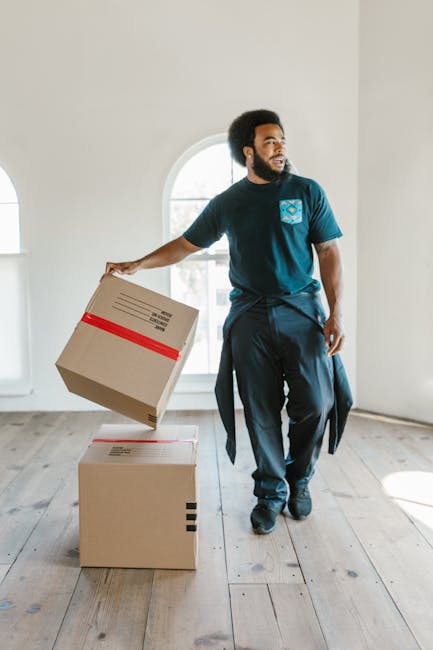 A man with a beard and dark curly hair, dressed in a blue t-shirt and dark trousers with a jacket tied around his waist, is inside a bright, neutral-colored room with large arched windows allowing natural light to fill the space. He is engaged in a home relocation task, lifting a medium-sized cardboard box wrapped with red tape, while standing next to two other boxes placed on a wooden floor. The boxes are made of brown cardboard, and one is stacked on top of the other. The room appears empty aside from the boxes, with clean walls and minimal decorations. The scene depicts furniture transport and packing during a house move, illustrating the process of loading items for transportation, which could be part of a professional removals service like those provided by Man with Van Battersea. The overall setting emphasizes careful handling of household items during a furniture relocation or home packing process leading to a move in Battersea, as highlighted by the reference to the Battersea removals real cost guide.