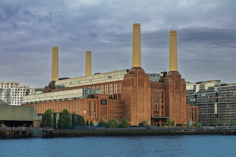 A large, historic industrial building with a reddish-brown brick facade and four tall, beige chimneys extending from the roof, situated along a waterfront. The building is surrounded by modern residential and commercial structures and has multiple windows and architectural details indicating its former industrial use. In the foreground, there is a section of water with a pier or dock on the left side, and the overall scene is under a cloudy sky. This setting illustrates the environment where professional house removals and furniture transport services by Man with Van Battersea are often coordinated, with careful loading of household items and furniture onto trucks for home relocation and moving logistics.