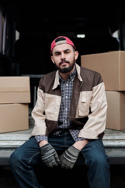 A man with a beard and glasses, wearing a white polo shirt and cap, stands outside a moving van during a home relocation process on Queenstown Road, Battersea, at sunset. He is in the process of loadi