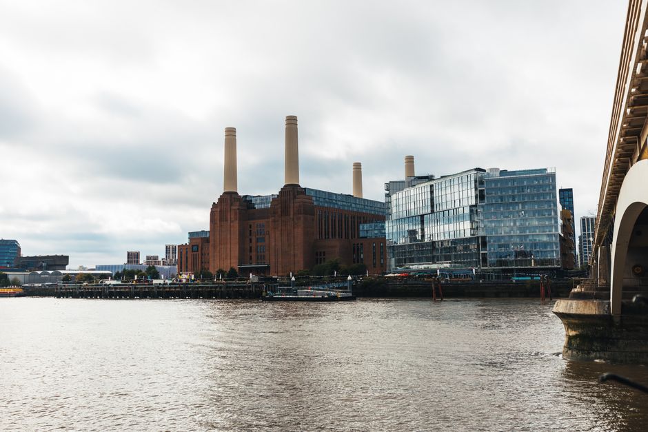 A large red-brick power station with four tall, cream-colored chimneys positioned against a cloudy sky. The building features vertical window strips and a modern glass extension on the right side. In